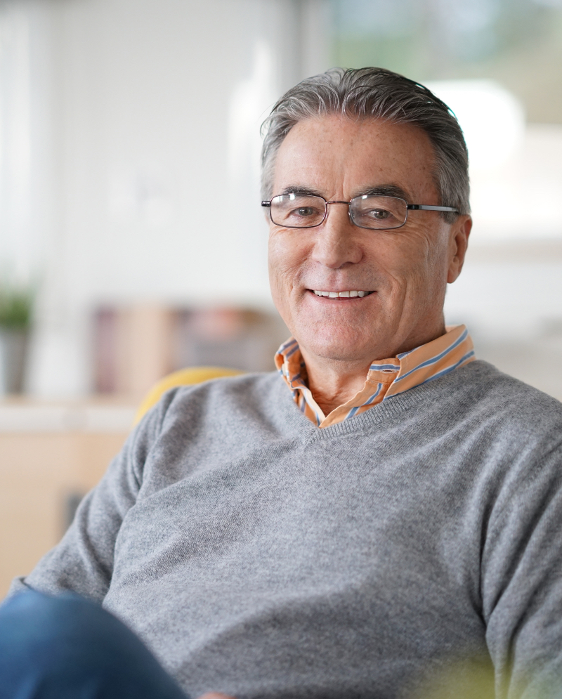 Older man wearing a gray v-neck sweater, sitting on the couch in his living room